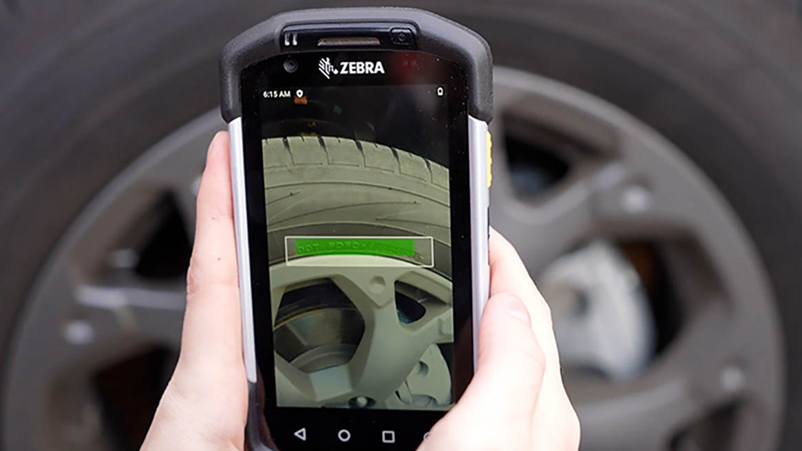 A person uses a Zebra handheld device to scan the tread on a car tire, utilizing Optical Character Recognition (OCR) technology to analyze and convert the tread patterns into machine-readable text for accurate documentation and analysis.